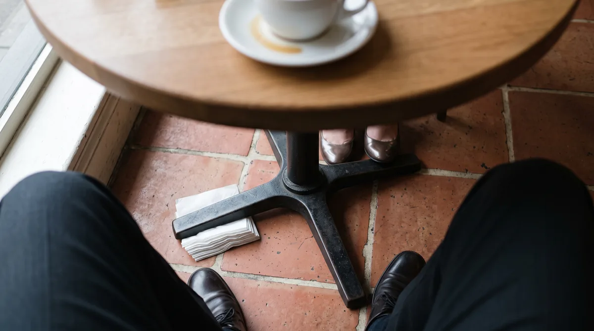 Looking down under a café table: a folded wad of napkins under one foot of its cast-iron base, coffee spilled into the saucer above, business shoes on both sides of the terracotta floor